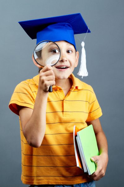 Portrait of curious boy in graduation hat looking through magnifying glass